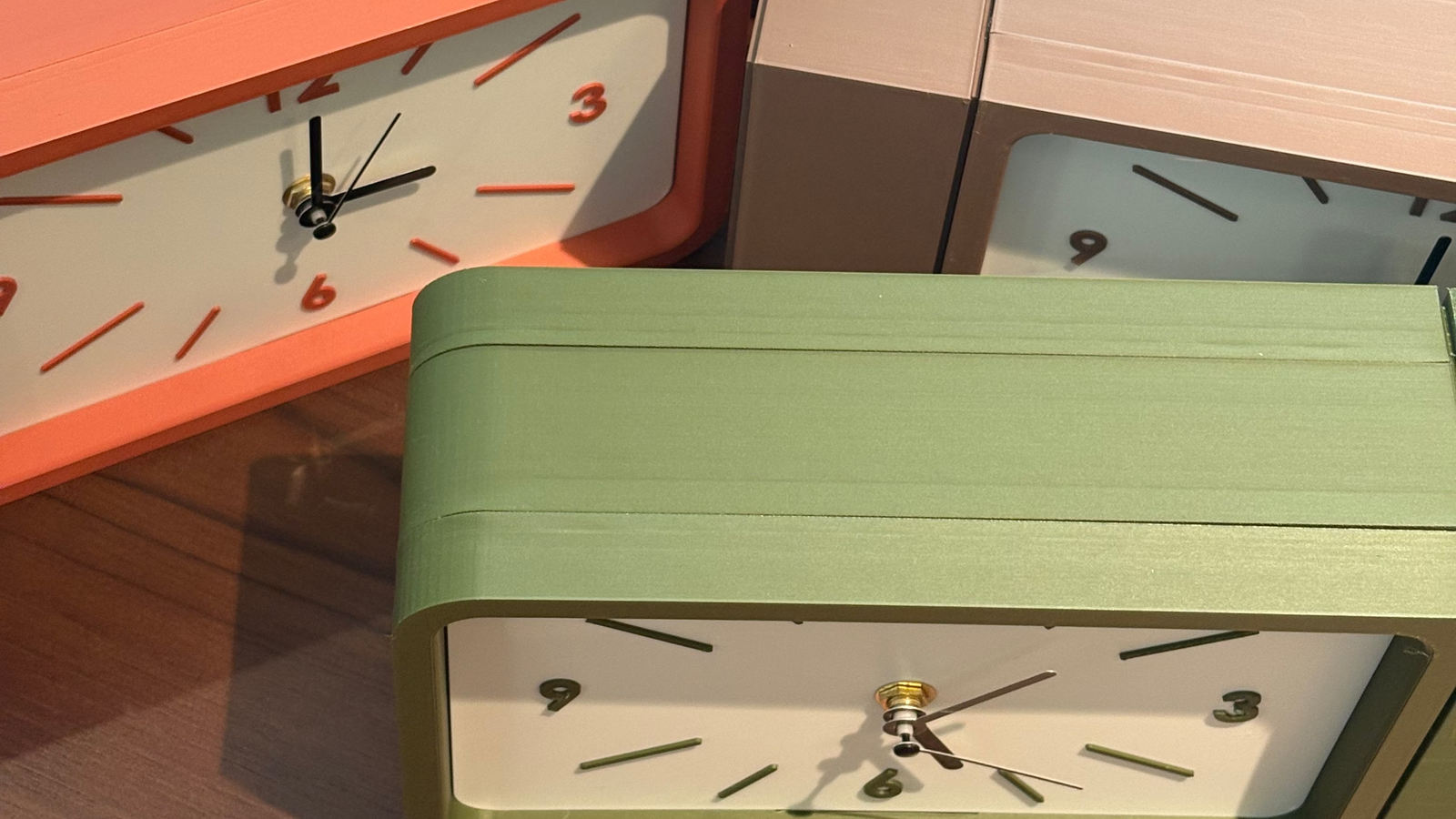 Four minimalist rectangular analog wall clocks with white faces and black numerals, shown in orange, sage green, beige, and black, arranged on a wooden table.
