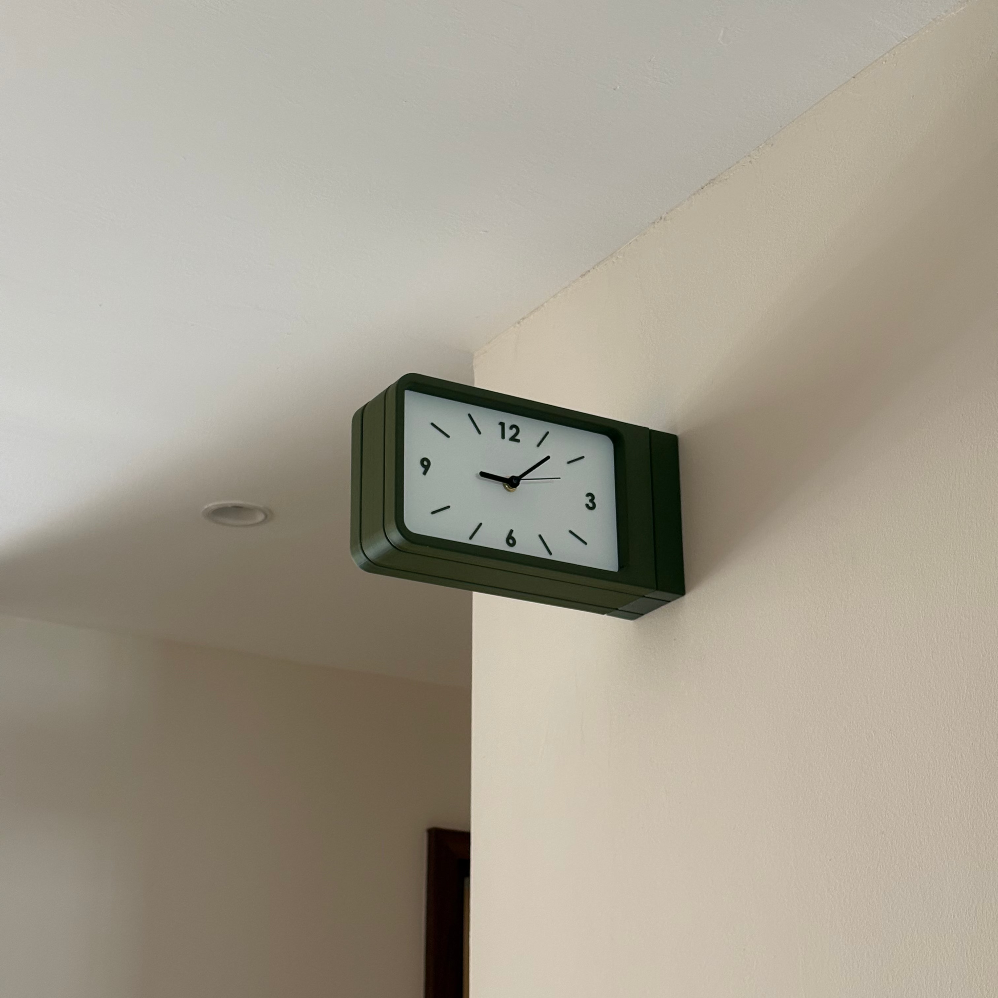 A rectangular wall clock with a dark green frame and a white face, mounted on a beige wall in an indoor room setting.