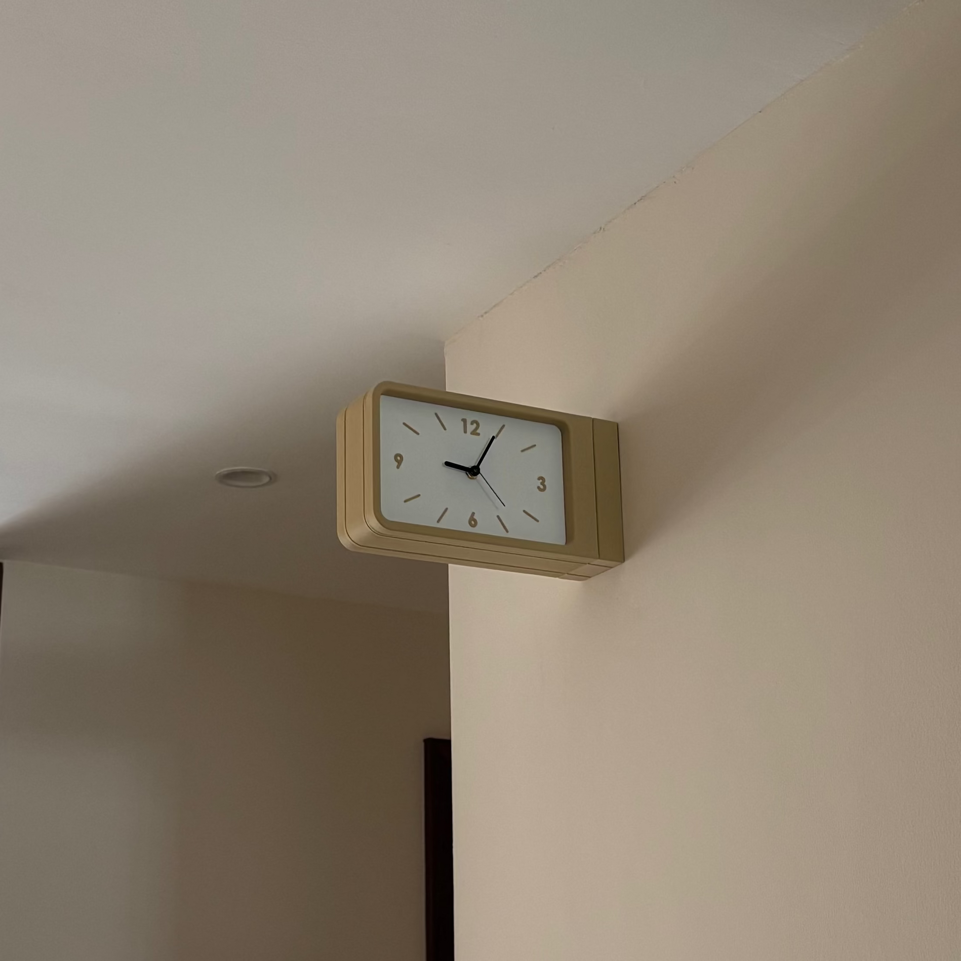 A rectangular wall clock with a light wood frame and white face mounted on a beige wall in an indoor hallway.