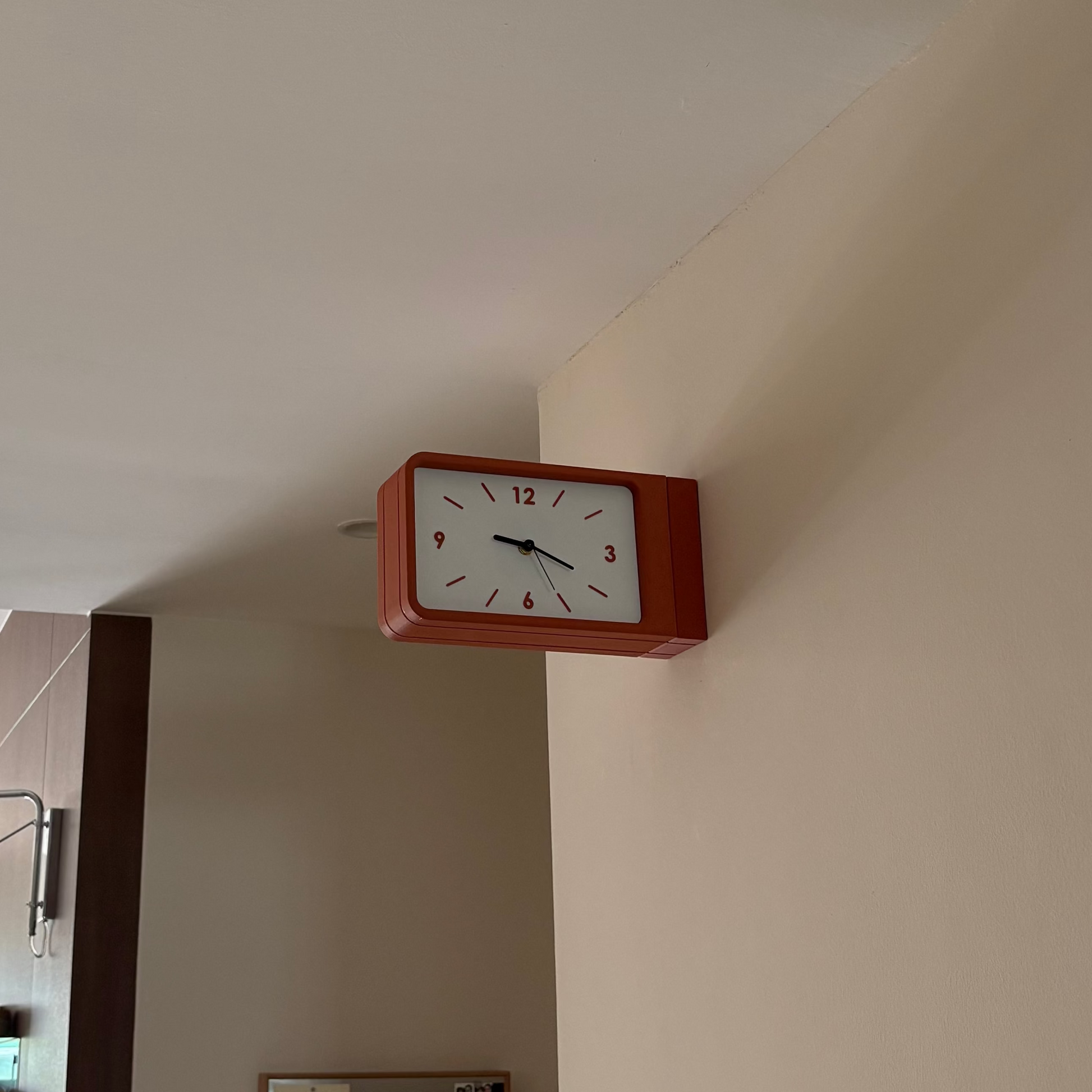 A rectangular wall clock with a reddish-brown frame and a white face, mounted on a beige wall in an indoor room setting.