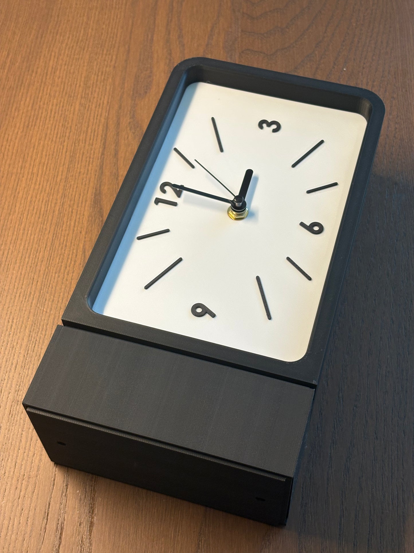 A modern rectangular wall clock with a matte black metal base and frame, a white dial face, and black hands, photographed on a wooden tabletop.