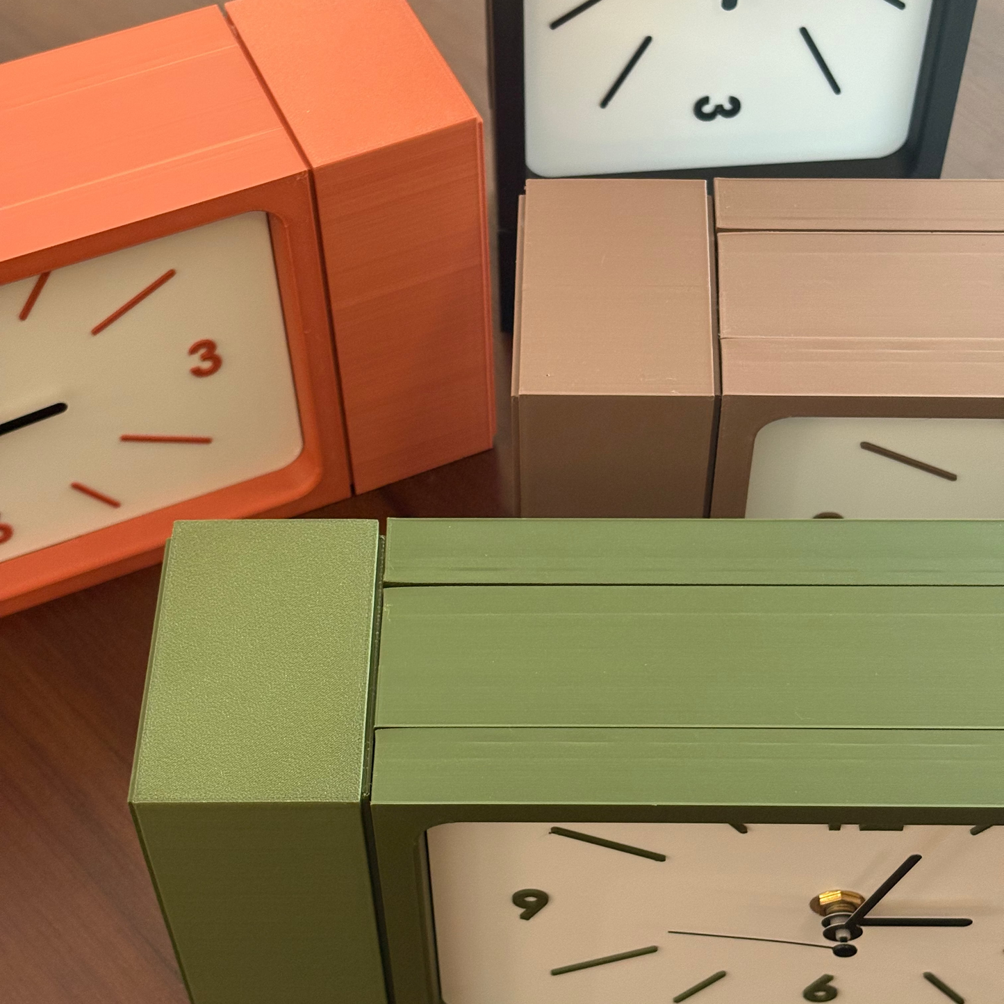Four retro-style rectangular desk clocks with minimalist white faces and black numerals, shown in orange, sage green, and beige tones on a wooden surface.