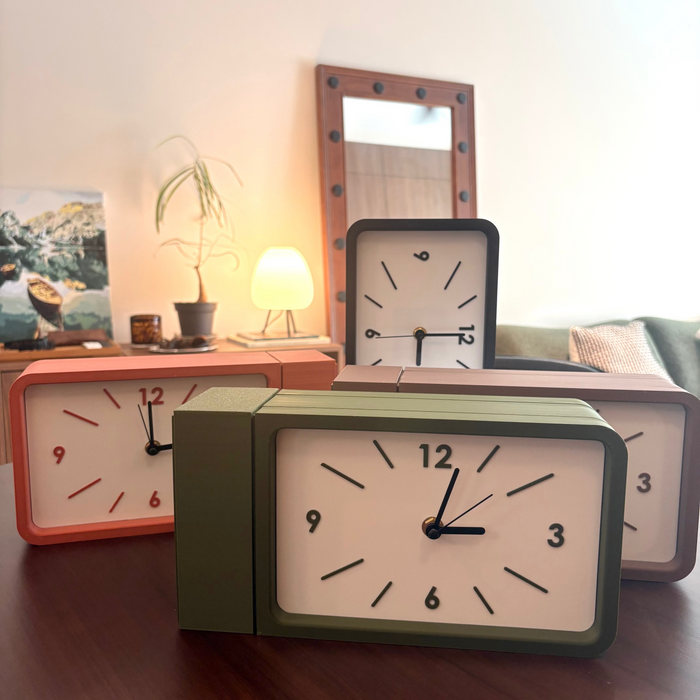 Four retro-style rectangular desk clocks with cream faces and black numerals, shown in green, coral, black, and beige frames on a wooden table in a living room setting.