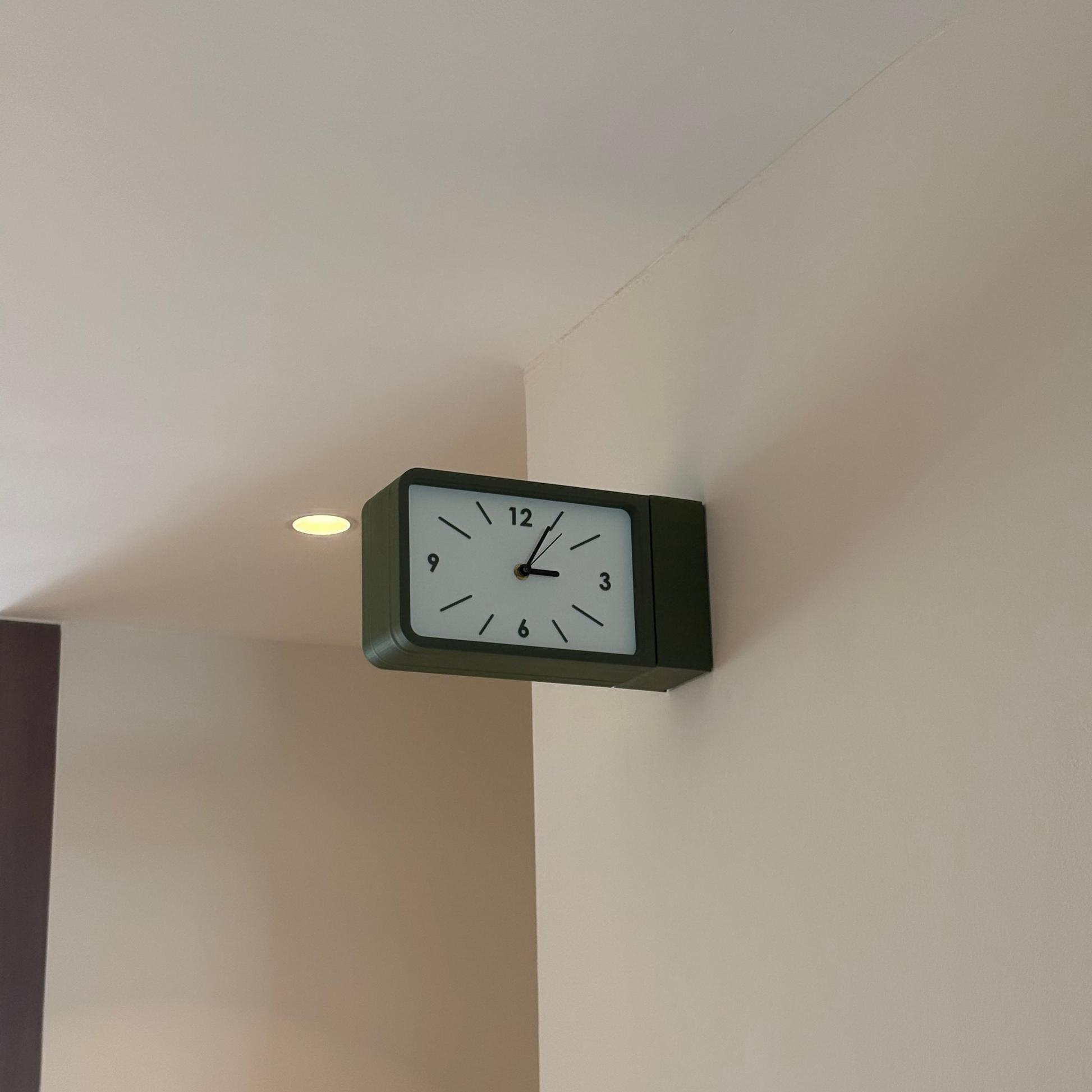 A rectangular wall clock with a dark green frame and white face, mounted on a beige wall in an indoor room setting.