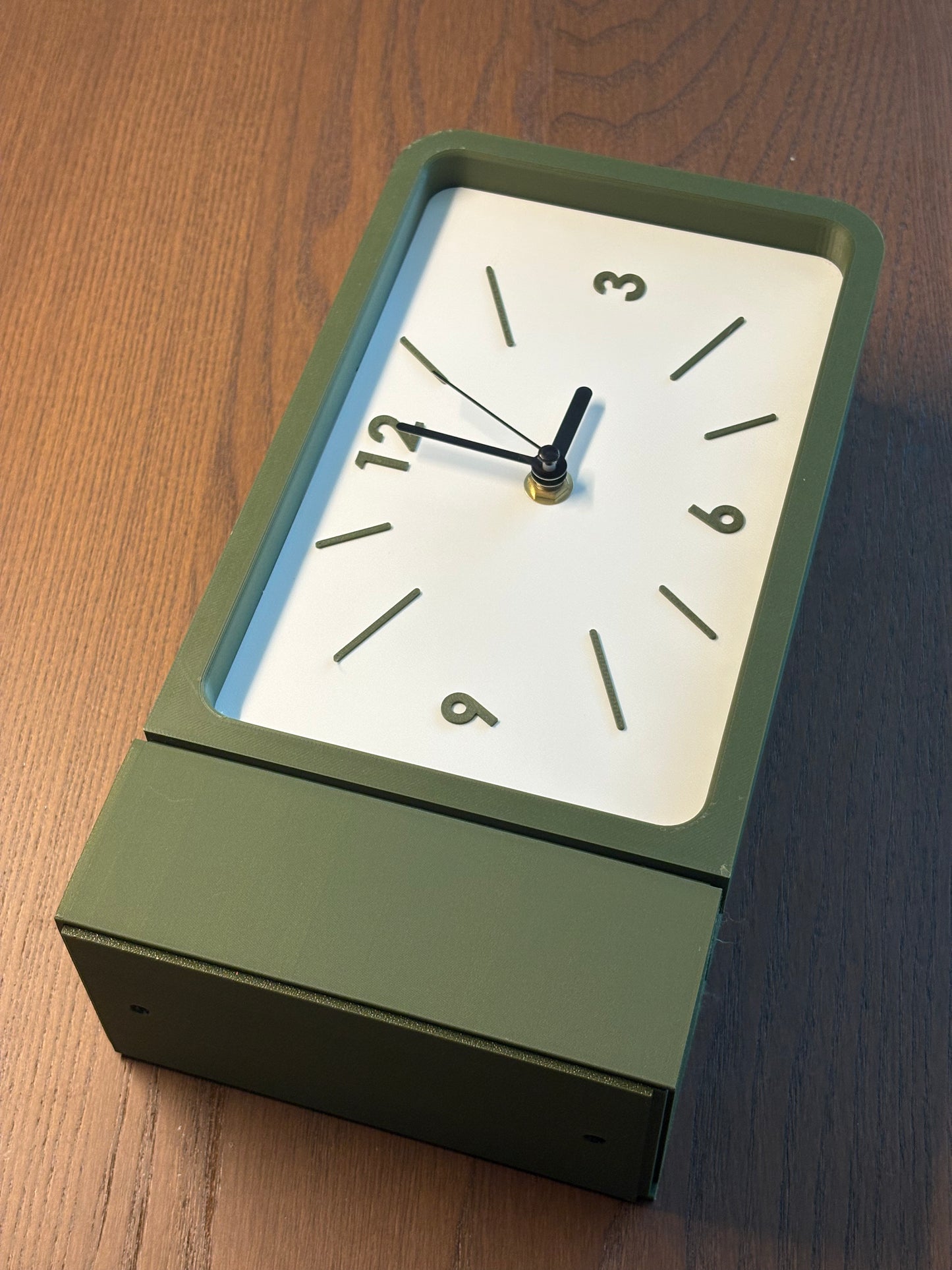 A rectangular desk clock with an olive green metal body and a white face, shown on a wooden tabletop.