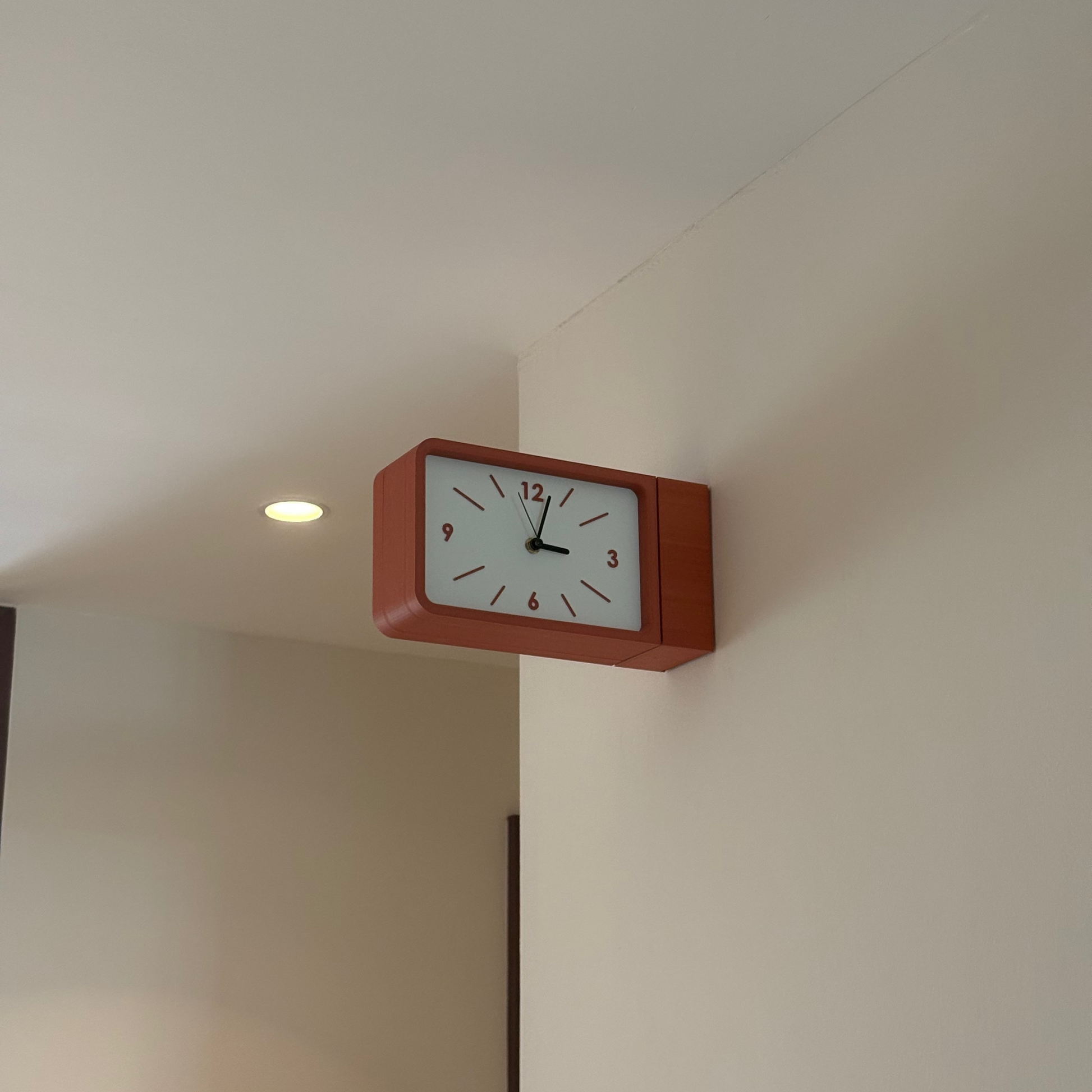 A rectangular wall clock with a reddish-brown wooden frame and a white face, mounted on an off-white wall in a real interior setting.