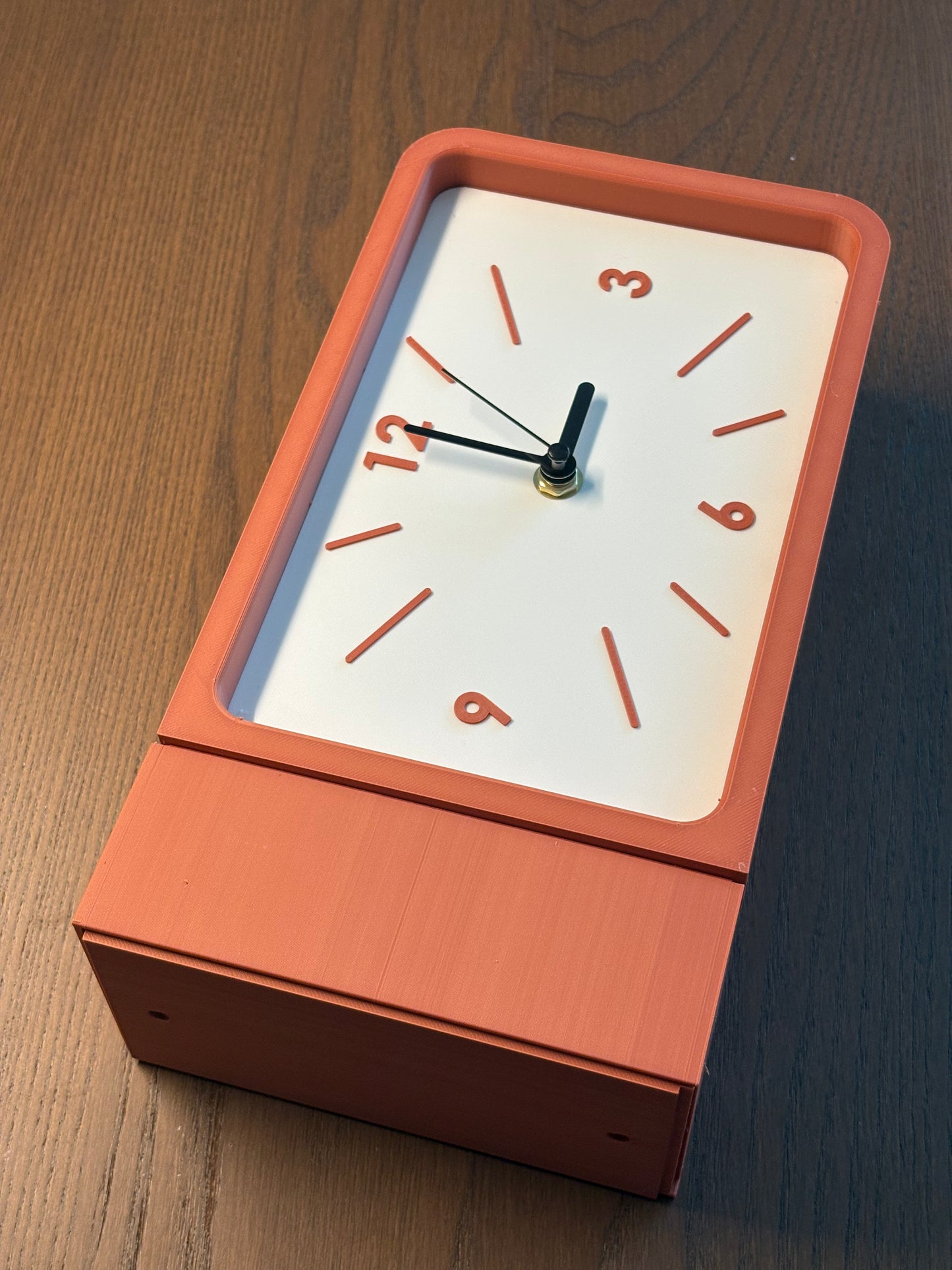 A rectangular analog wall clock with a matte terracotta orange frame and a white face, shown on a dark wood tabletop.