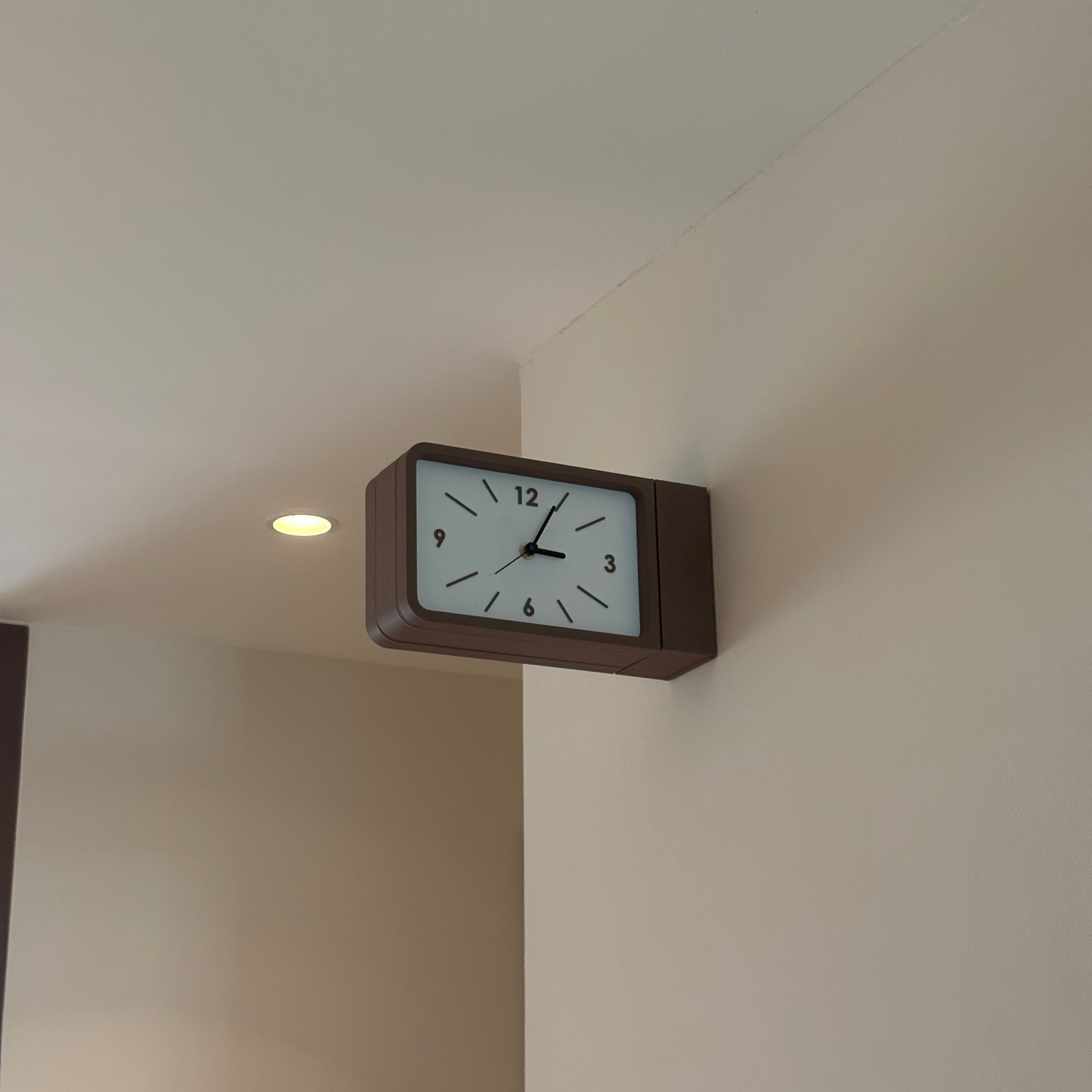 A rectangular wall clock with a dark brown frame and a white face, mounted on a beige wall in an indoor setting.