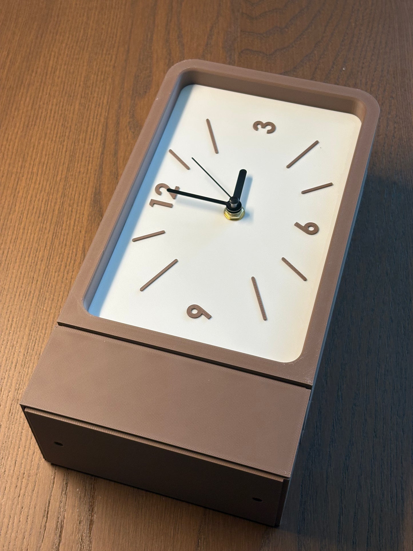 A modern rectangular wall clock with a matte taupe-brown frame and a white face, shown resting on a dark wood tabletop.